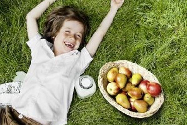 Ein lachendes Mädchen in weißer Bluse liegt auf der grünen Wiese, neben sich eine Flasche Milch und ein Korb Äpfel