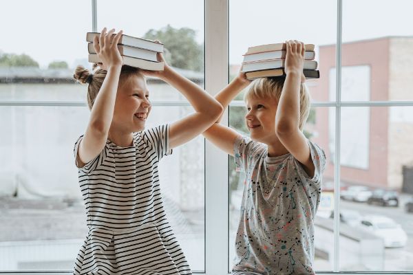 Zwei Kinder sitzen lachend am Fenster und balancieren Bücher auf ihren Köpfen