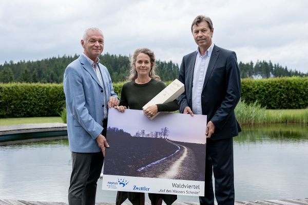 Die Preisträgerin Regina Hügli steht mit Rudolf Damberger von Zwettler und Generalsekretär Günter Liebel auf einem Steg, sie hält den Neptun-Preis und einen Druck ihrer Fotografie in der Hand, im Hintergrund sind Wasser und Gebüsch zu sehen
