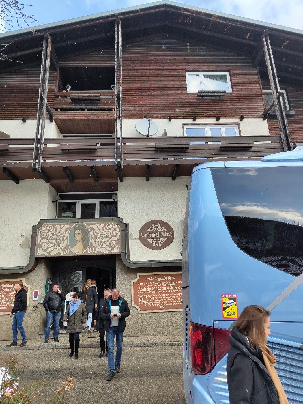 Personen verlassen ein traditionelles Gasthaus zur Straße hin. Rechts steht ein Bus.