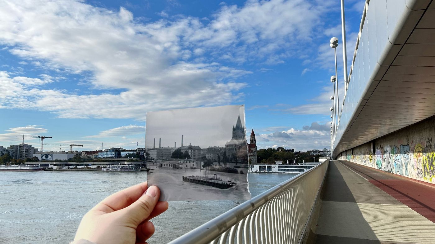 Ein Foto auf einer Donaubrücke in Wien zum Ufer, wo eine Hand ein altes Foto desselben Abschnittes hält.