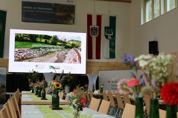 Hintergrund Foto von Ortschaft, seitlich Oesterreichfahne und Niederoesterreichfahne, Besprechungstisch laengst angeordnet, rechts und links Stuehle und der Tisch geschmueckt mit mehreren Vasen mit Blumen 