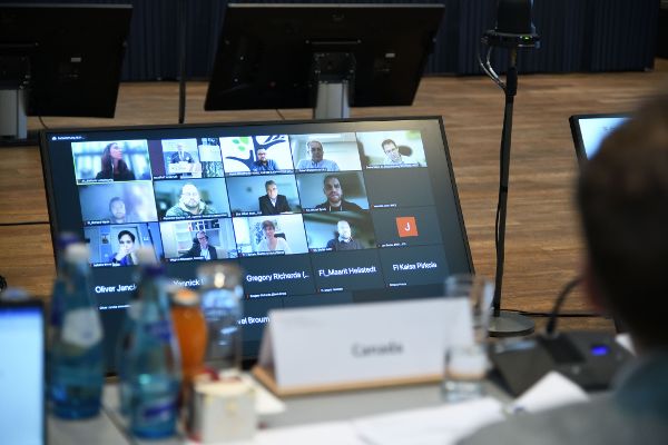 Man blickt über den Rücken eines Teilnehmers und den Tisch hinweg auf den großen Bildschirm. Hier sind die Teilnehmerinnen und Teilnehmer des Zoom-Meetings zu sehen. 