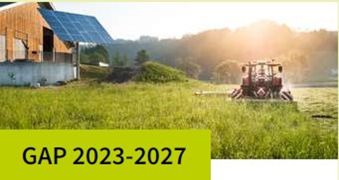 GAP 2023-2027. Das Foto zeigt einen Traktor mit Mähwerk, der eine grüne Wiese neben einem landwirtschaftlichen Gebäude mit Photovoltaik-Anlage mäht.