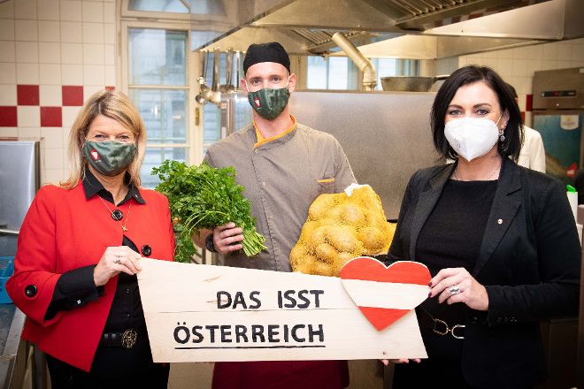 Bundesministerinnen Köstinger und Tanner in einer Heereskantine gemeinsam mit einem Heeresbediensteten, halten regionale Lebensmittel und ein Schild "Das isst Österreich"