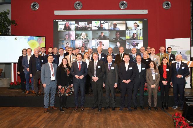 Gruppenfoto der Teilnehmer:innen an der woodPoP-Veranstaltung inklusive der Online-Teilnehmer im Hintergrund auf einer LED-Wand. Landwirtschaftsminister Totschnig steht zentral in der ersten Reihe. 